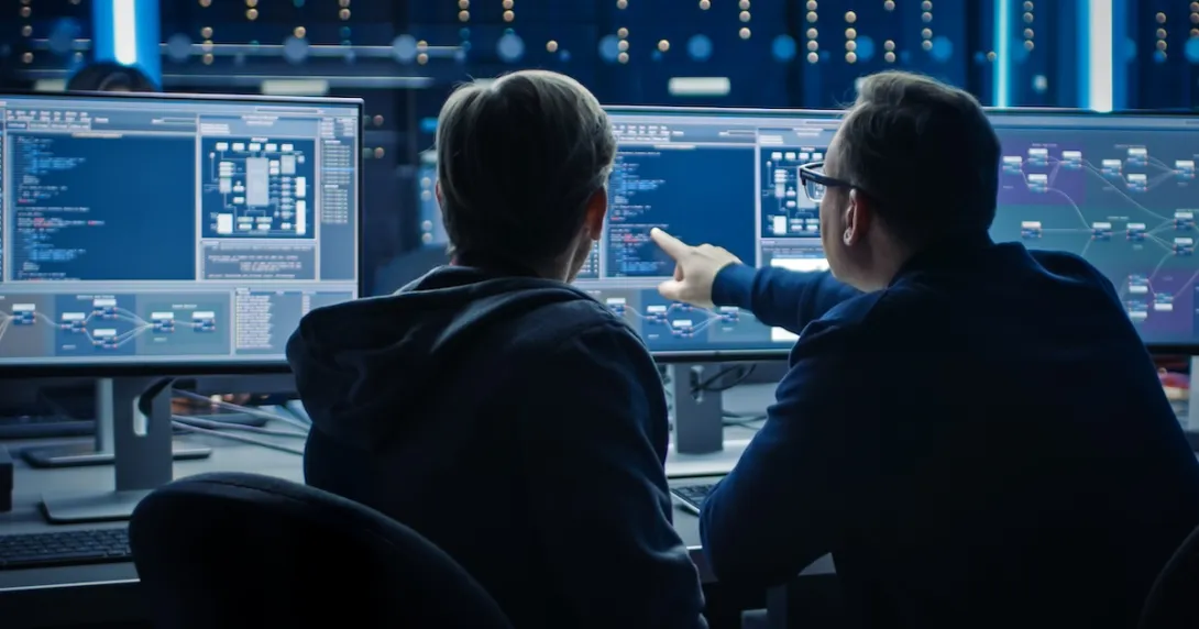 Two people sitting in front of three computer monitors analyzing data on the screen
