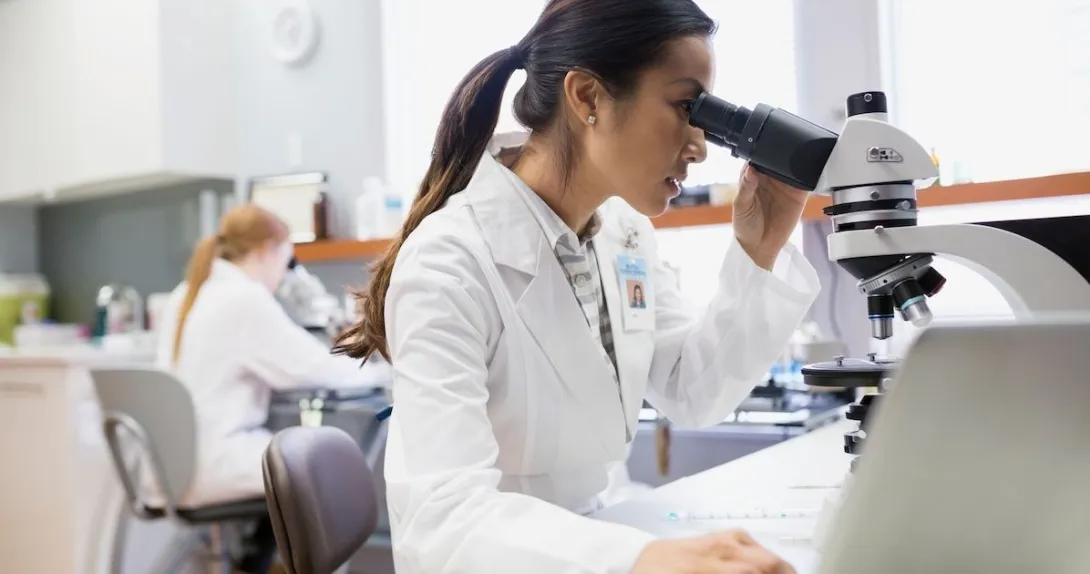
  Person sitting down looking into a microscope while wearing a lab coat
